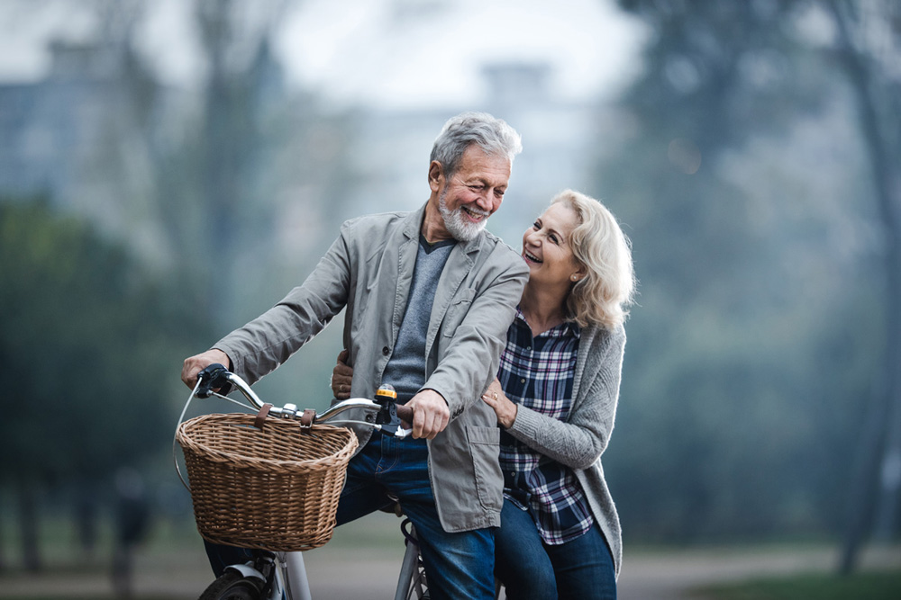 Happy mature couple communicating while riding on a bike in the park. Copy space.